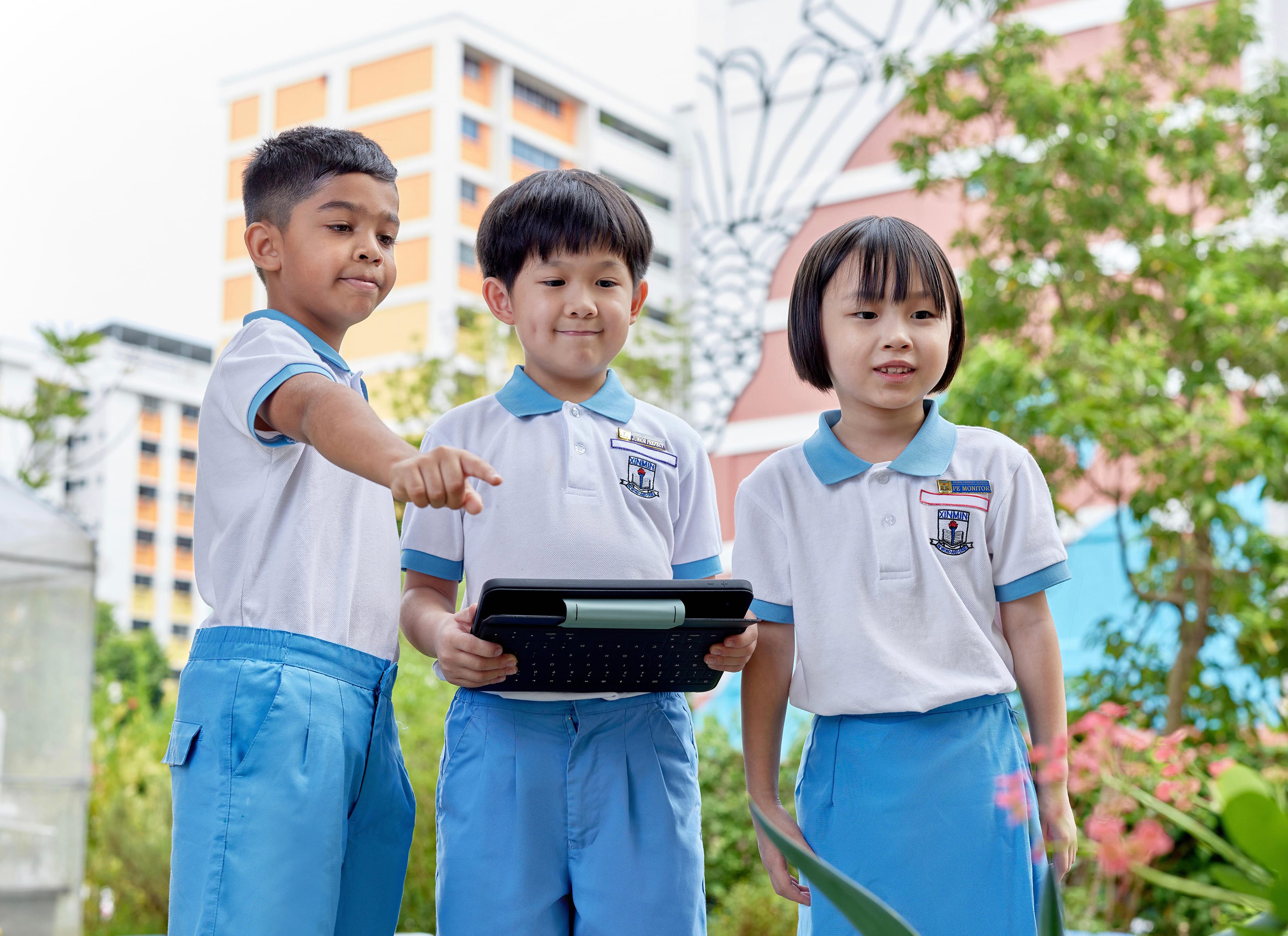 Three schoolchildren in white uniforms outdoors; one points while another holds a tablet keyboard.